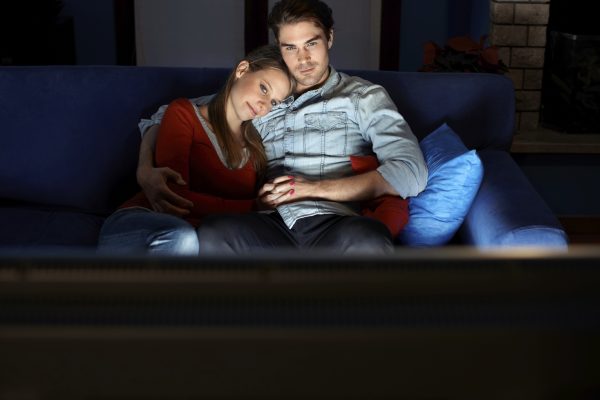 young heterosexual couple hugging on sofa and watching movie on tv at home. Horizontal shape, front view, copy space