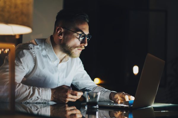 Bearded young businessman working on modern loft office at night. Man using contemporary notebook texting message, holding smartphone, blurred background.