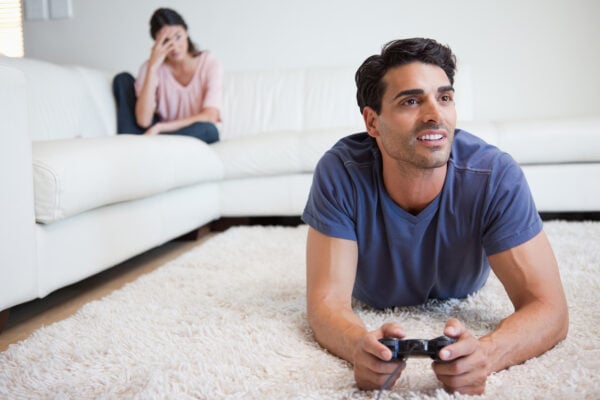 Man playing video games while his girlfriend is getting mad at him in their living room