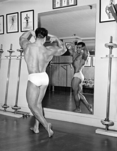 1955: Body-builder Leo Robert, holder of the Mr Universe title, flexing his muscles while looking at his reflection in a large mirror in a gymnasium. (Photo by Alan Oxley/BIPs/Getty Images)