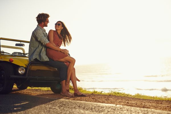 Young couple looking into each other's eyes. Romantic young couple sitting on hood of their car enjoying the moment outdoors with copy space. Young couple in love on a road trip.