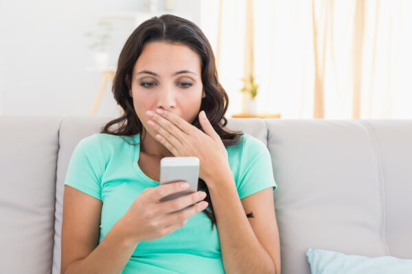 Shocked brunette texting on the phone at home in the living room